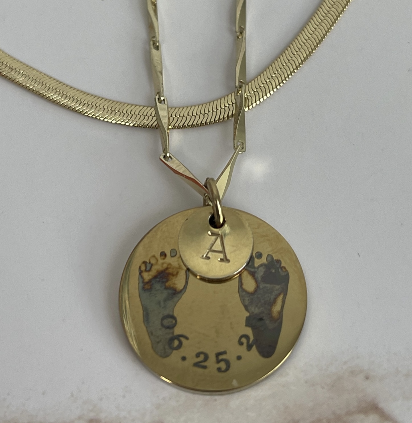 Gold round pendant engraved with baby footprints, a date, and a smaller initial charm